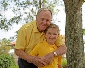 A heartwarming photo of golf legend Jack Nicklaus in a yellow polo shirt, hugging a young boy also wearing a yellow polo, standing outdoors near a tree.