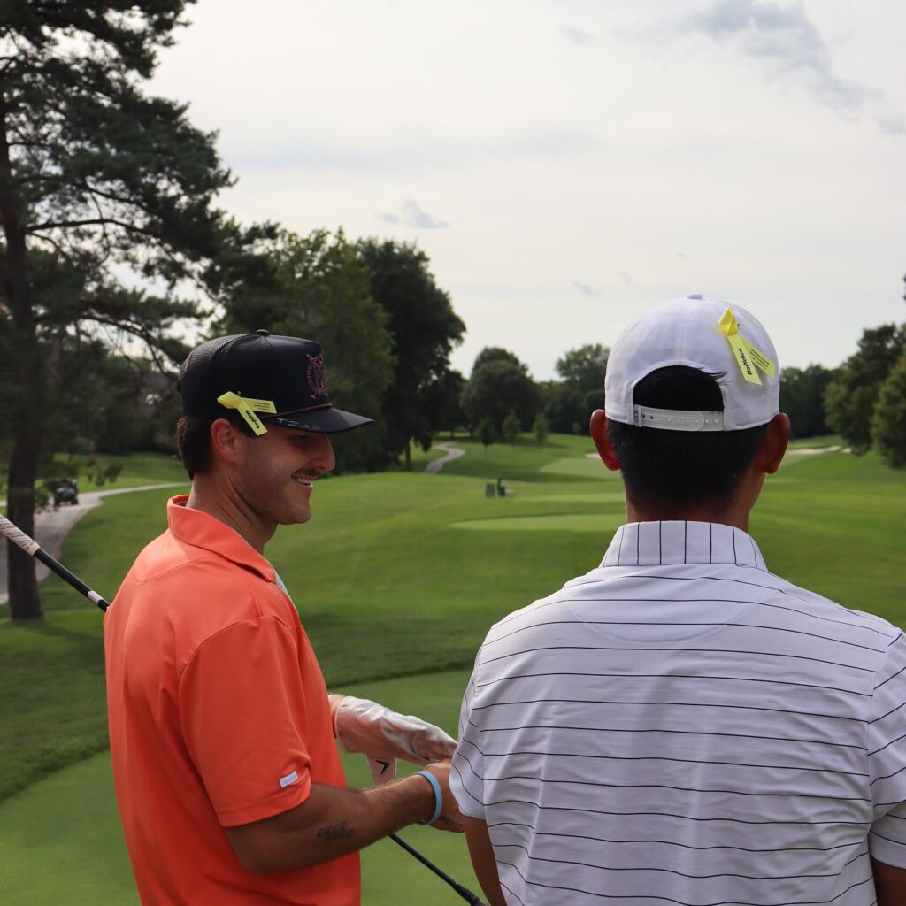 Two golfers on a green course seen from behind; the golfer on the left wears a black hat and the golfer on the right wears a white Nike hat, both adorned with yellow Play Yellow campaign ribbons.