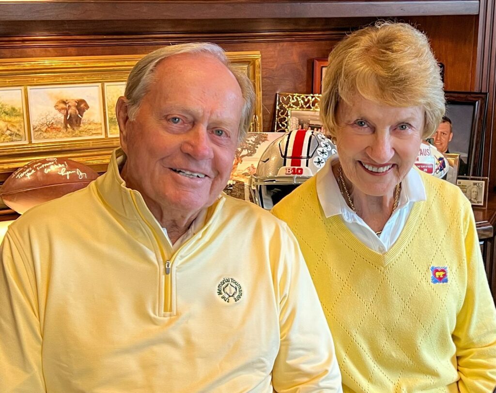Golf legend Jack Nicklaus and his wife Barbara Nicklaus smiling together indoors. Jack wears a pale yellow quarter-zip with The Memorial Tournament logo, and Barbara wears a yellow diamond-pattern sweater featuring a small Golden Bear heart logo.