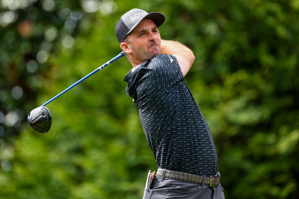 Professional golfer Denny McCarthy wearing a grey hat and a patterned black polo, captured in the middle of a powerful driver swing on a lush green course.
