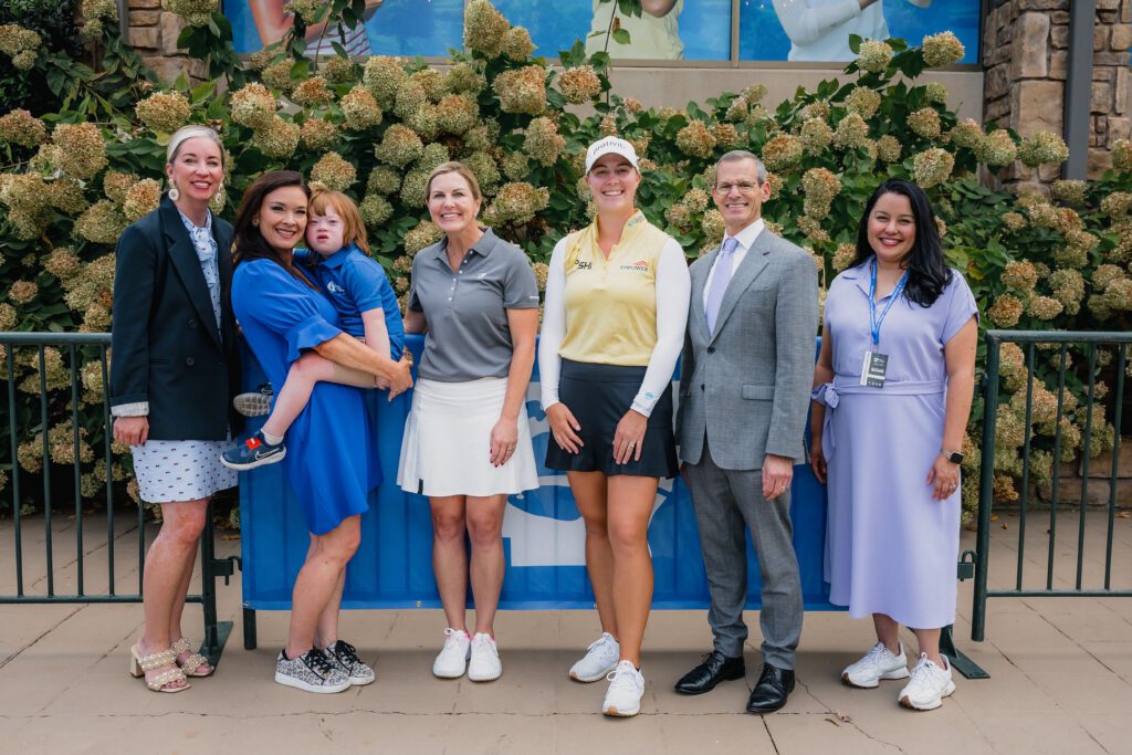 A group of seven people, including professional golfer Alexa Pano in a yellow and black golf outfit, pose together outdoors in front of a blue banner and a lush green hedge.