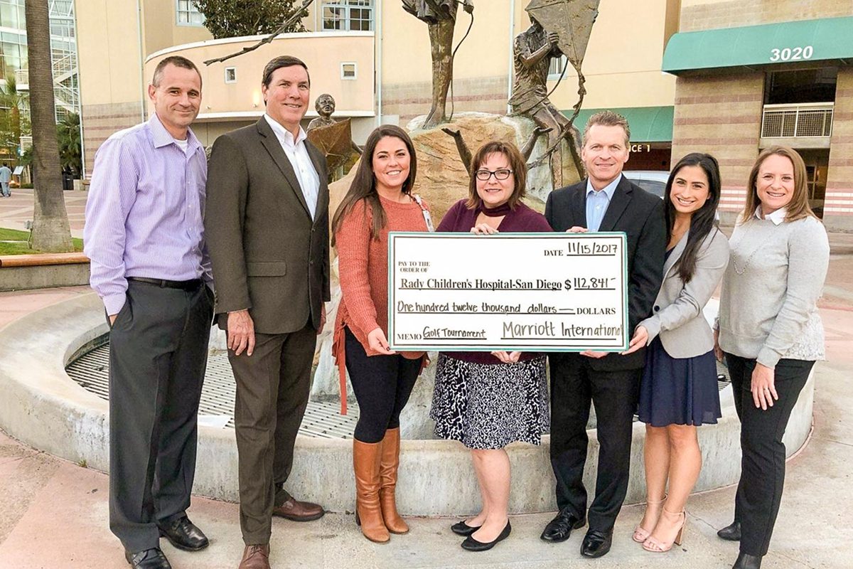 : A group of seven people posing in front of a fountain, holding a large ceremonial check from Marriott International for $112,841 made out to Rady Children's Hospital-San Diego