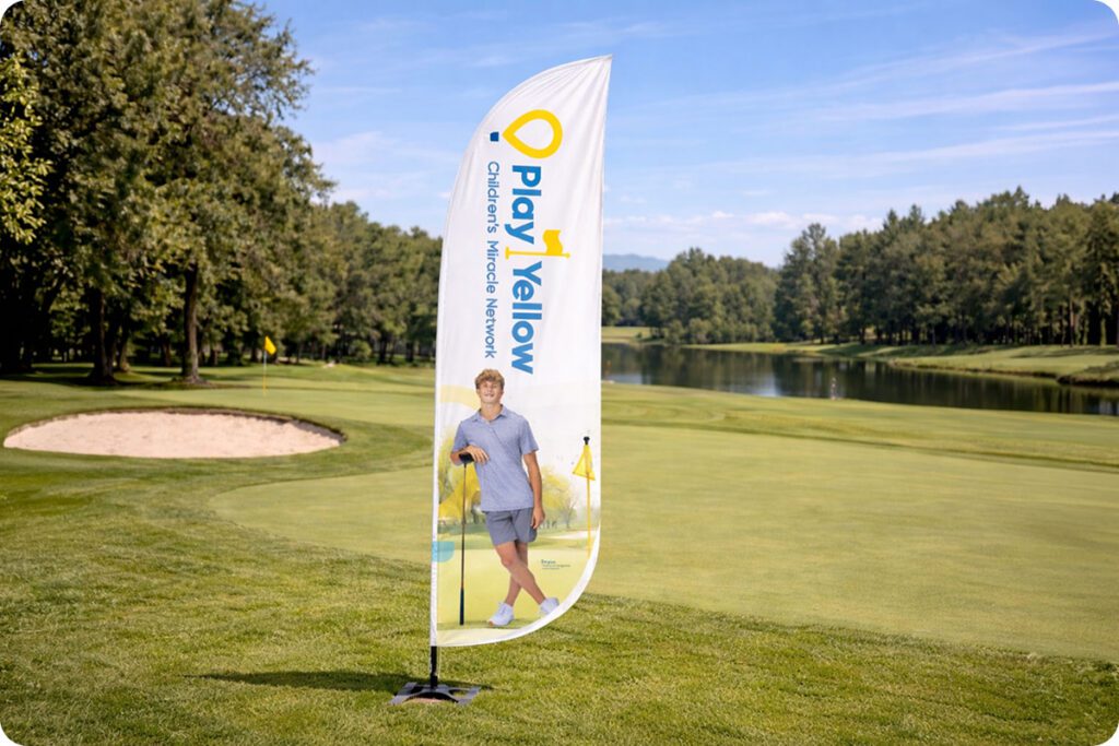 A white Play Yellow feather banner featuring a young golfer and the Children's Miracle Network logo stands on the edge of a golf course fairway near a lake.