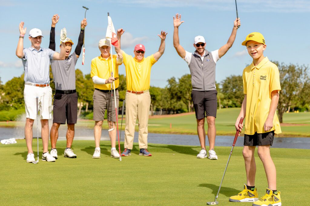 A young boy in a yellow Play Yellow polo and Golden Bear hat putts on a green while a group of five men, including several wearing Play Yellow gear, cheer with their hands raised in the background.